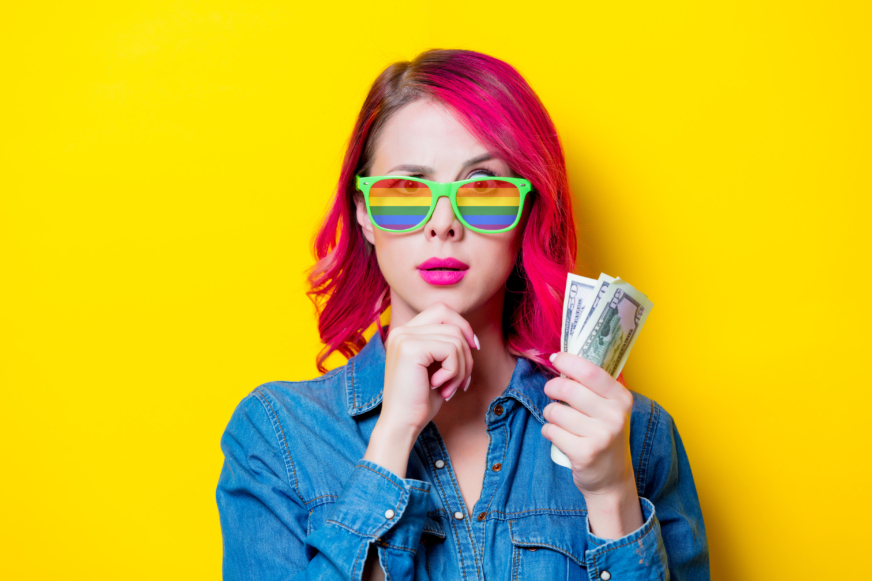 A young woman with bright red hair stands smiling against a yellow background.  She wears rainbow colored sunglasses and holds a wad of cash while looking pensive.