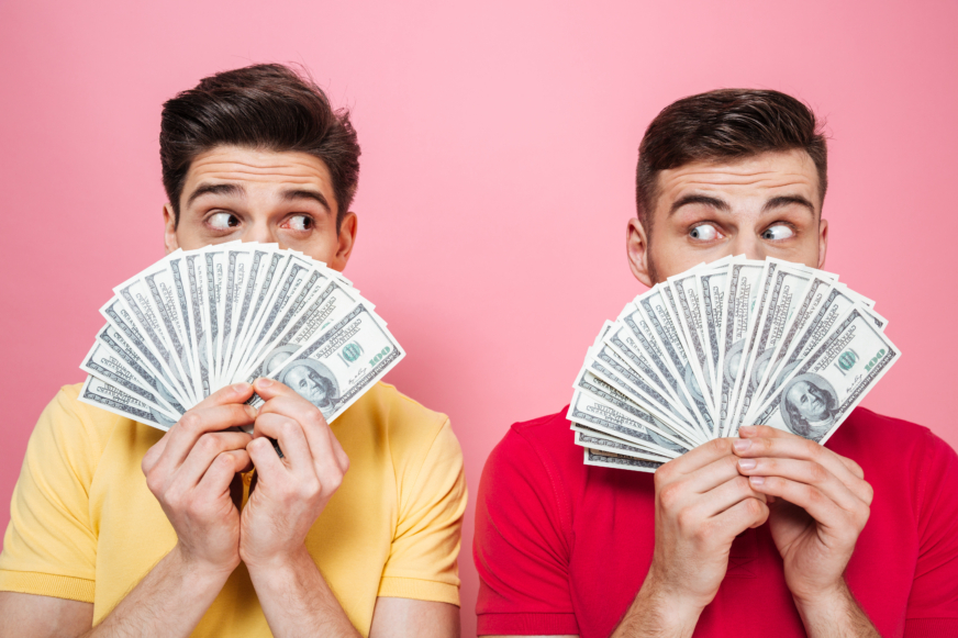 Two gay men stand in front of a pink background holding $100 bills in front of their open faces.